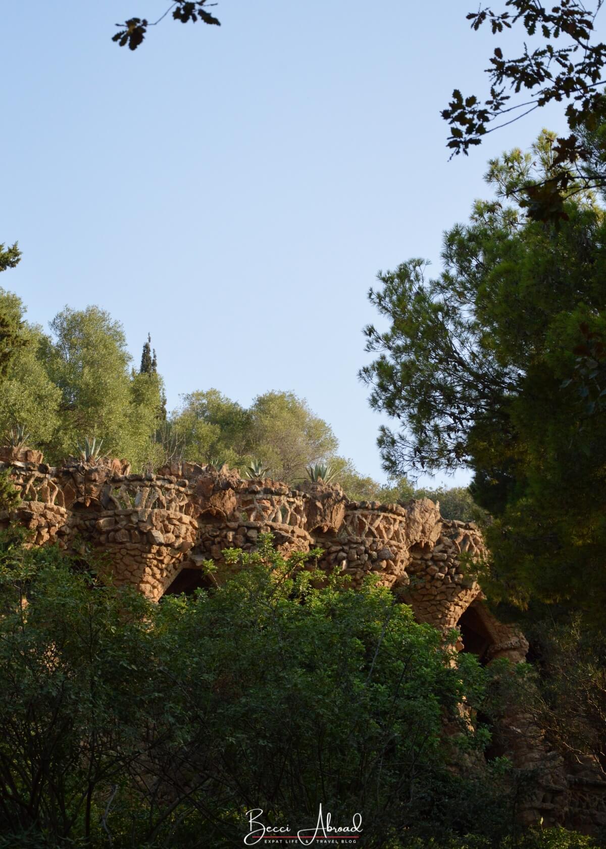 A pathway winding through Park Güell in Barcelona, bordered by lush greenery and Gaudí’s stone viaducts blending into the natural landscape.